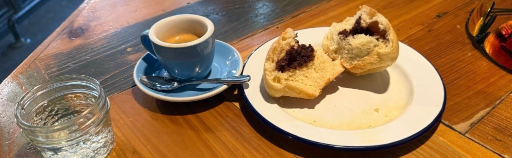 A photo showing a shot of espresso in a blue porcelain cup, a sidecar of sparkling water and a brioche sourdough bun broken open to show a chocolate center.